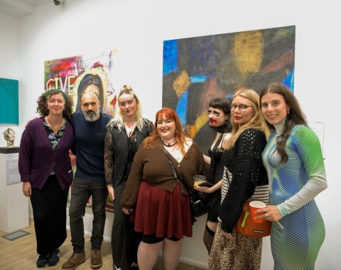A group of students - six women and one man - stand in a line in front of a wall of art work.